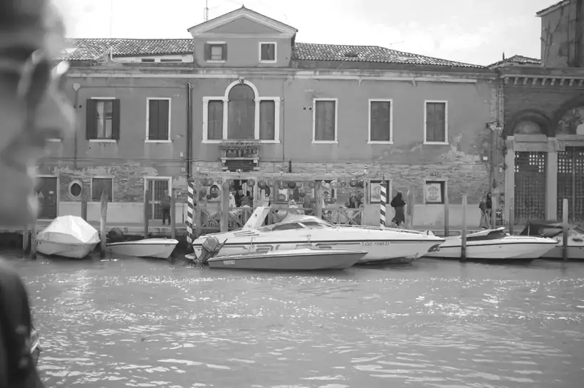 Barche a motore ormeggiate in un canale davanti a un palazzo intonacato, sagoma sfocata in primo piano, bianco e nero