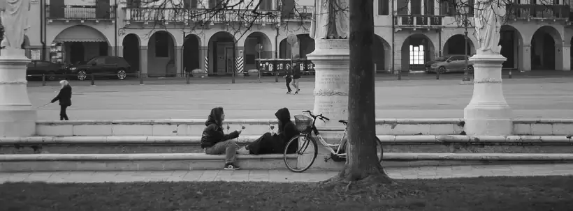Due ragazzi seduti sui gradini di una piazza con statue e portici, bici appoggiata e tronco in primo piano, bianco e nero
