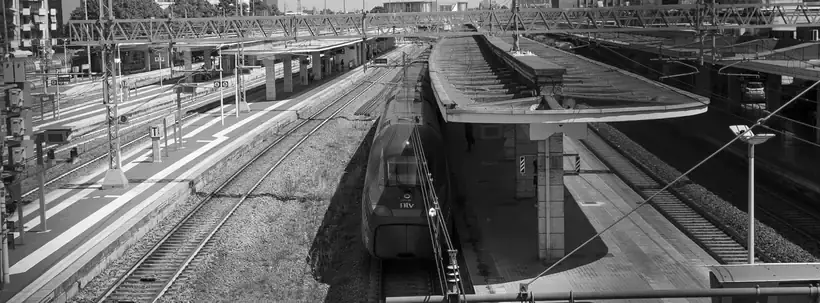 Dall'alto sopra i binari, il muso scuro di un treno fermo tra pensiline curve e cavi aerei, bianco e nero
